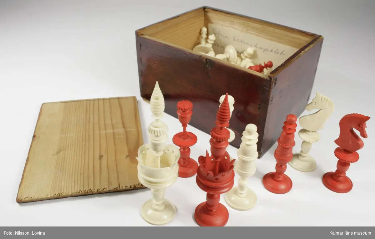 KLM 28703:13. Schackpjäs. Komplett uppsättning schackpjäser bestående av 32 pjäser, 16 vita och 16 röda. Dessa förvaras i mörkbrun ask av trä med skjutlock. Några av pjäserna är skadade.
