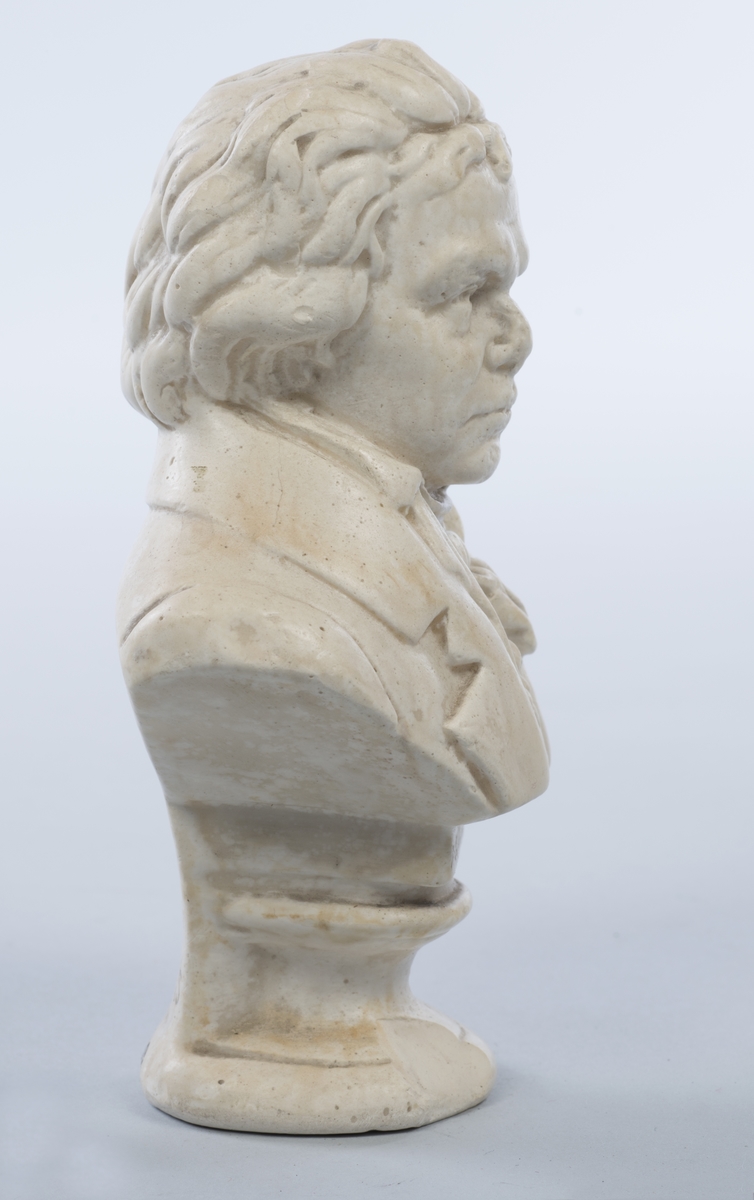 Beethoven iført åpen frakk, vest, skjorte med høy krage og knyttet tørkle i halsen. Rund sokkel.