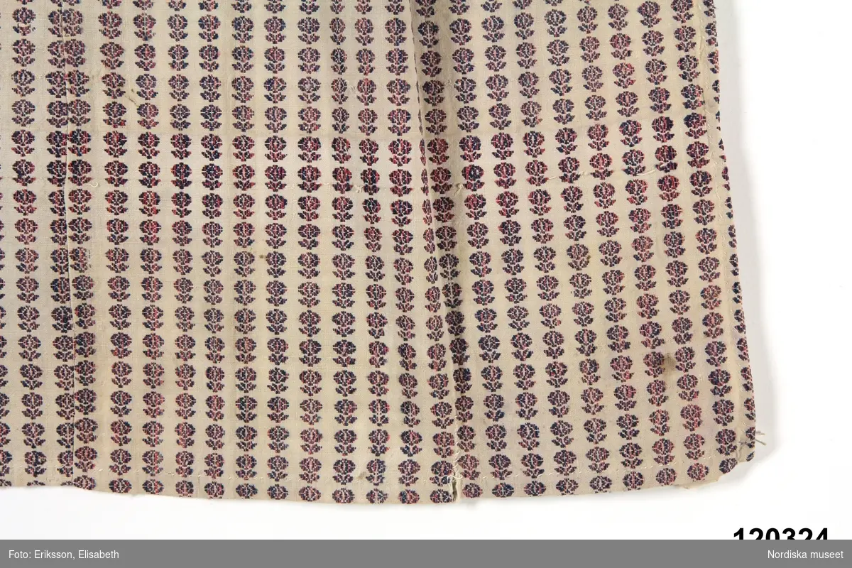 Förkläde i bomull med flerskaftad mönstervävning i blått och rosarött på vit botten. Täta bårder med små stiliserade  blommönster där blått och rött blandats. 
Mönstervävningen påminner mycket om småmönstrade vävnader moderna under 1820-talet som finns i den tidens vävprovböcker. 
Förklädet sytt av 3 hopskarvade  längder med  handsydda fållar på 3 sidor och har  dragsko i midjan med iträdd lingarnssnodd.
Anm. Blekt och slitet med småhål speciellt på vänster sida
Dessa bladförkläden vävdes sannolikt av speciella väverskor.  Jfr med förkläde 137.669 soml enligt uppgift skall vara tillverkat 1840.
/Berit Eldvik 2012-03-26