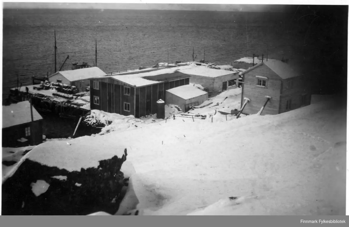Bildet tatt i Øksfjord under gjenreinsninga.På kaia kan man se et fiskermottak, eid av Njord Handels og Industri A/S. På bygningene kan man se tak og vindu. Man kan se at det ligger masse snø på bakken, i bakgrunnen kan man se havet.