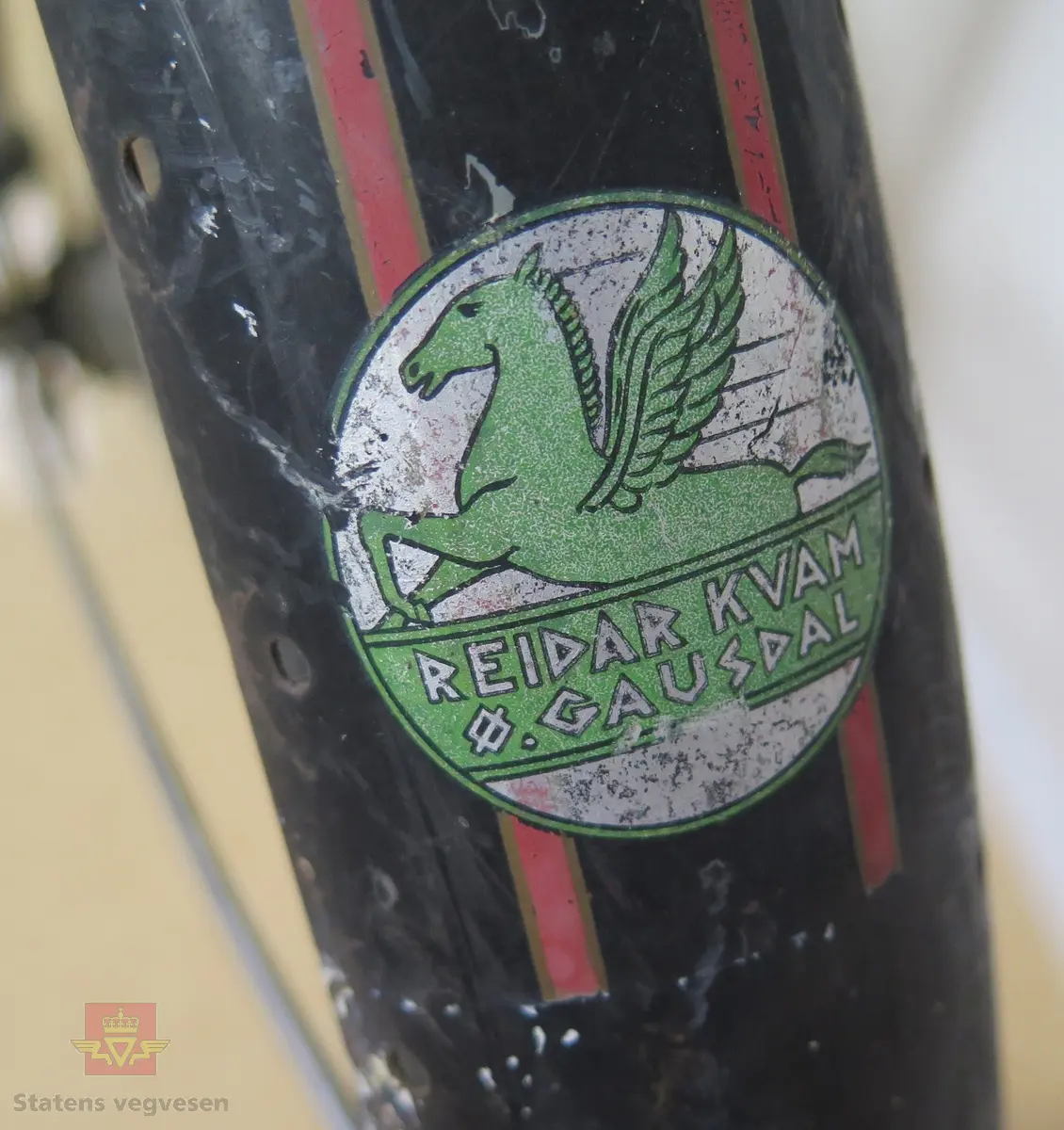 Tråsykkel, damemodell. Har to hjul, styre, sete, pedaler, frontlykt, ringeklokke, bagasjebrett, dynamo og refleks bak. Svart med rød staffering. Brunt sete og røde holker på styret. Sykkelen er i hovedsak lagd av metall, gummi og skinn.
Emblem med teksten: REIDAR KVAM Ø. GAUSDAL MIL.