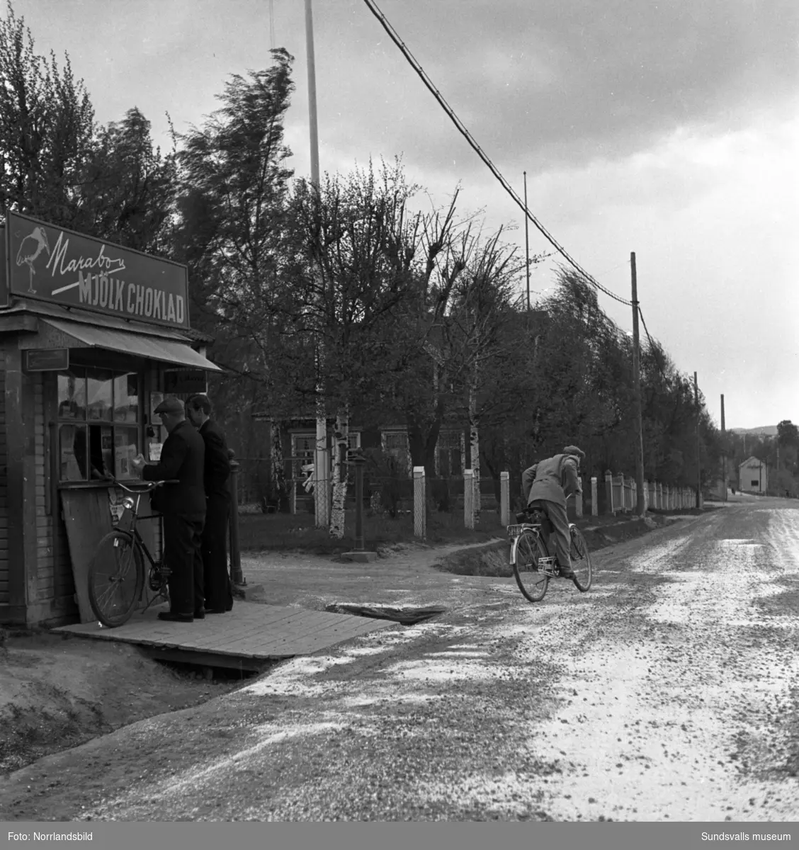 Två män handlar i en liten kiosk invid vägen i Stadsmon. En annan man ger sig iväg på sin cykel.