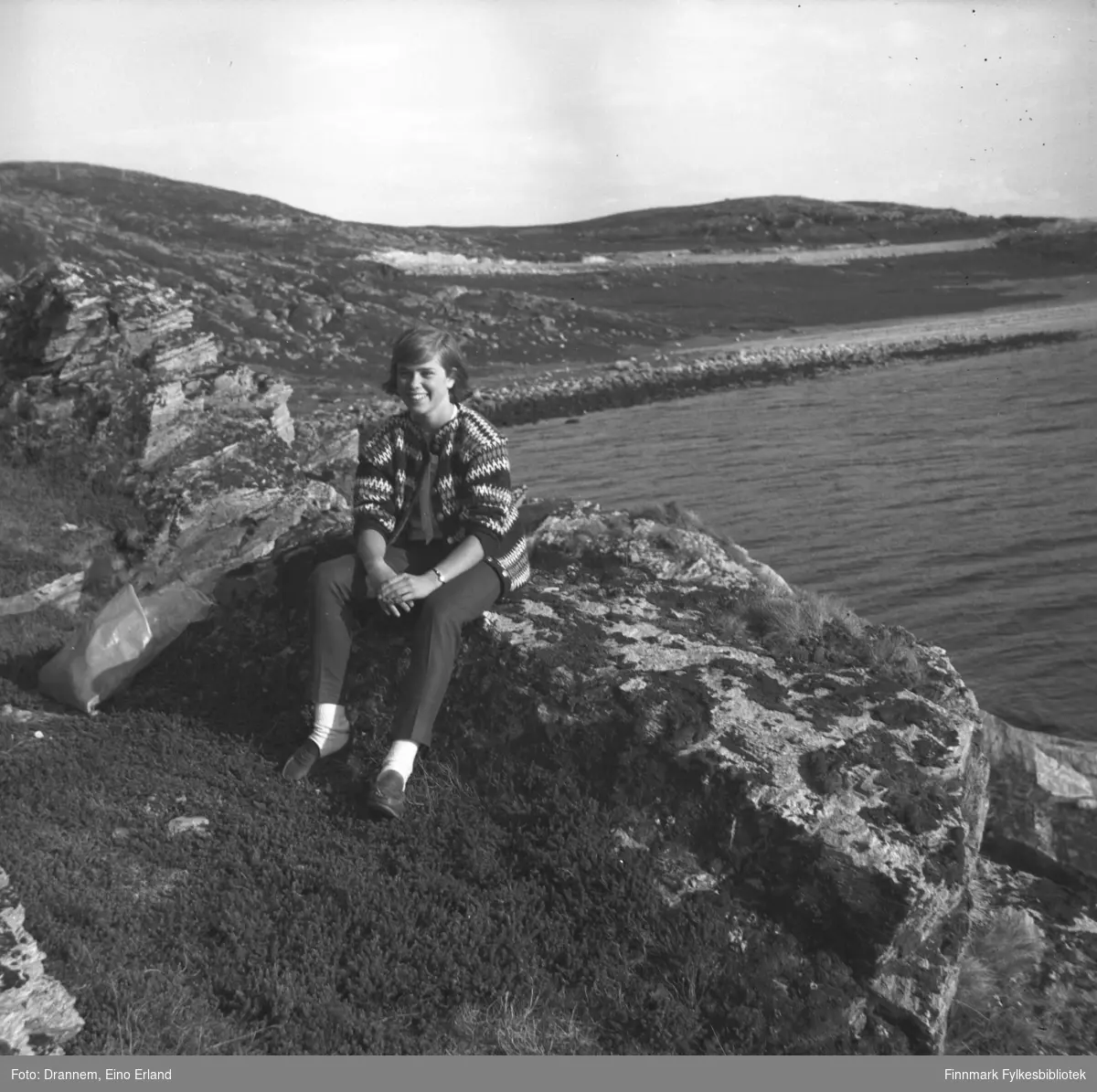 Marit Gabrielsen sitter på en stein i veikanten til Rv94 med Kargenes i bakgrunnen.