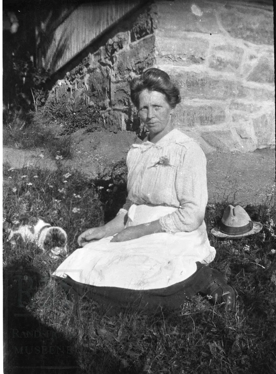 Dame sitter i gresset, en katt ligger ved siden av henne og en hatt som tilhører Marius Aasheim. Damen er Lise Aasheim.