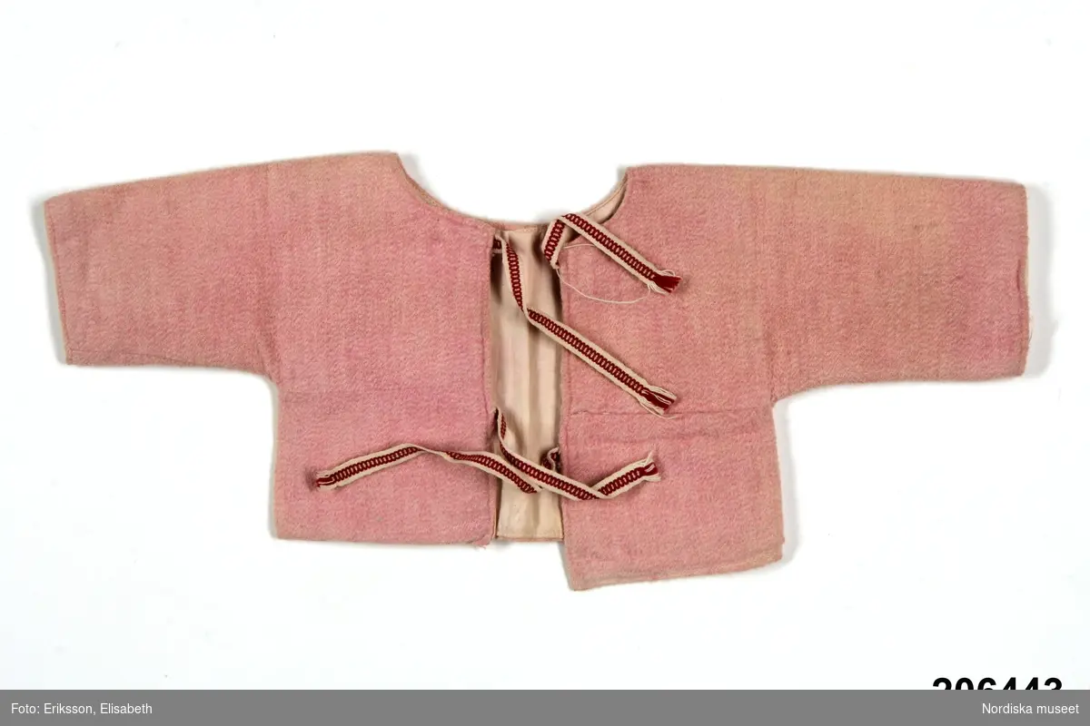 Spädbarnströja  av rosa bomullsflanell, ett bålstycke med sömmar över bröstet, öppen fram med två knytband  i vitt med rött mönster, Fodrad med vit bomullslärft.  Sydd på maskin.
Se även 206 441-446.
/Berit Eldvik 2013-02-07