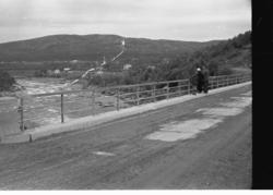 Tre personer står på Neiden bru og ser på elva. Bebyggelse i