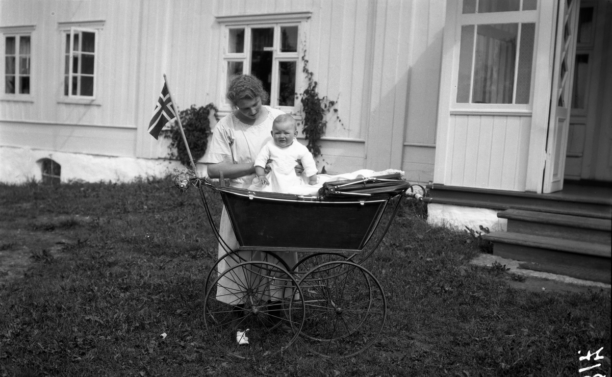 Uidentifisert kvinne med barn i barnevogn foran et stort våningshus. På barnevogna er det festet et norsk flagg.