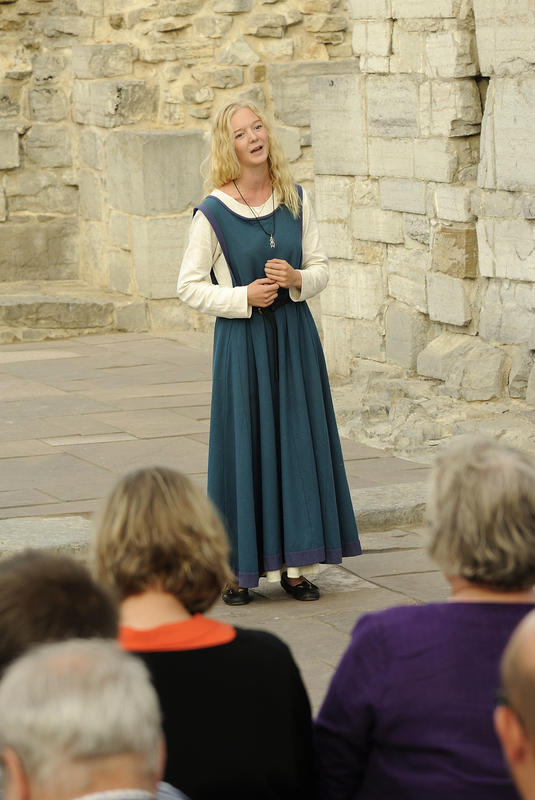 A young woman with long, blond hair and a blue Medieval dress is standing and singing in front of an audience in the cathedral ruins.