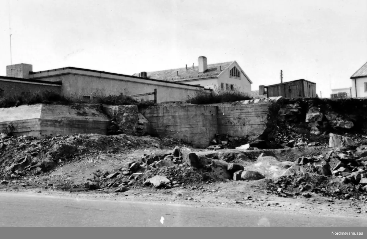 Siste rest av fjellhaugen der Arbeiderforeningen og Rådstua stod. I bakgrunnen ser vi rester av en tysk bunker som ble bygget under krigen 1940-45, og over denne bunkeren hadde da Arbeiderforeningen stått før bybrannen i de sene aprildagene 1940. Bak denne igjen ser vi det nye bygg for folkebiblioteket. I bakgrunnen ser vi Indremisjonsbygget og oppover Marstrands gate. Badet (seinere Nor-Pro) til høyre, revet (først parkering for ny-banken, pr 2007 boligblokk). Her holdt lastebilsentralen til i 1960-åra, pr 2007 Hydro bensinstasjon, og drosjene er fortsatt pr 2007 foran Misjonshuset. 
Inngangen støpt igjen i januar 2017 i forb. med butikk i Tinghuset. ( Regelbau 633 for M19 bombekaster) (info fra Bjørn Ove Angvik - 30.01.2017)
 

Nordmøre Museums fotosamling