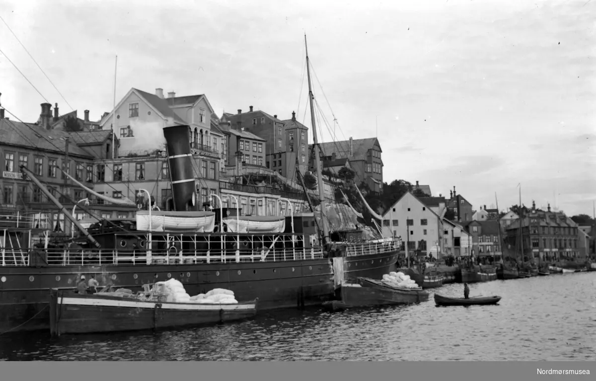 Et bilde fotografert ytterst fra Piren på Kirkelandet i Kristiansund, hvor vi ser flere fartøyer fortøyd langs Vågeveien, med blant annet en hurtigrute samt flere fiskebåter. Fra Nordmøre Museums fotosamlinger.