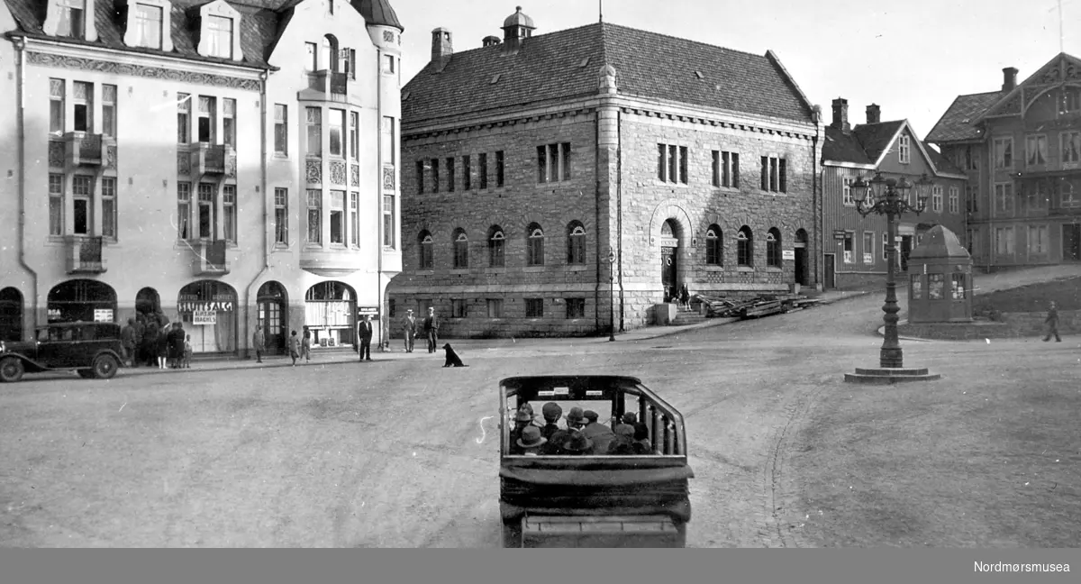 Torvet/Torget i Kristiansund, 
-Astrid Giertsen Modes hadde sluttsalg juni 1933, så bildet må være tatt rund den perioden.(info: Stein Ryther)  
-mennesker, bil, bygninger, bank, hotell, vei, hund, kiosk, gatelys, lysmast, arbeiderforening, avislokale, trykkeri, butikker, rutebuss,.... Bildet er tatt fra hjørnet med Grand Hotell, som hadde flere butikker i første etasje i front mot torget, og kinolokale på baksiden. Merk at en av butikkene har ";sluttsalg";, ";opphørssalg";. med stor kø ved inngangen. Bernstorffstredet, med det nye bygget til Norges Bank, hvor tidligere Bernstorffskolen, senere sjømannskolen ble revet for å gi plass til banken. Deretter avisen Tidens Krav's lokaler med trykkeri og redaksjon. Så ser vi hjørnet av Arbeiderforeningen. Foran Arbeiderforeningen ligger en av byens mange Narvesen-kiosker, og foran den ser vi byens egenartede gatelys, bedre kjent som ";lysekrona"; som var ett av flere klengenavn. Tidligere hadde ";lysekrona"; fem kupler. 
Denne står i dag 2007 foran Festiviteten og er logo for Operaen i Kristiansund. I forgrunnen ser vi en åpen rutebuss. Leonard Hjelle kunne i 1936 starte opp ringbussrute i Kristiansund i 1936 da brua mellom Gomalandet og Nordlandet var ferdig og åpnet.
 Et stort fremskritt for kristiansunderne som inntil da hadde vært avhengige av båt for å kunne komme til Nordlandet.
 Fra Nordmøre Museum sin fotosamling.