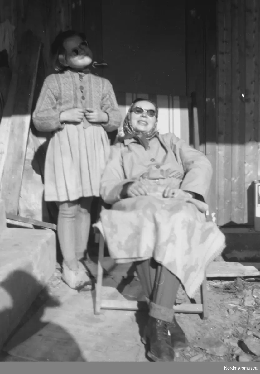 Foto av en kvinne sittende i en solstol, med en liten pike stående ved siden av. Trolig enten familie, slekt eller bekjente av fotografen, som sannsynligvis er John Myren. Datering er ukjent, men trolig mellom 1930 til 1960. Stedet er også ukjent, men kan være fra Sunndal kommune. Fra Nordmøre museums fotosamlinger. 

