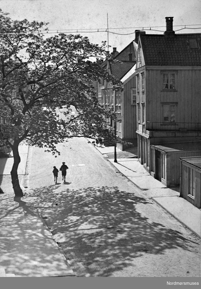 To gutter springer i Storgata.  Vi ser butikker, verksted og bolighus til høyre i bildet.  Telefon/strømledninger henger over gata.
Samme sted er fotografert etter bombingen av Kristiansund i aprildagene 1940. (se KMb-045.0002)