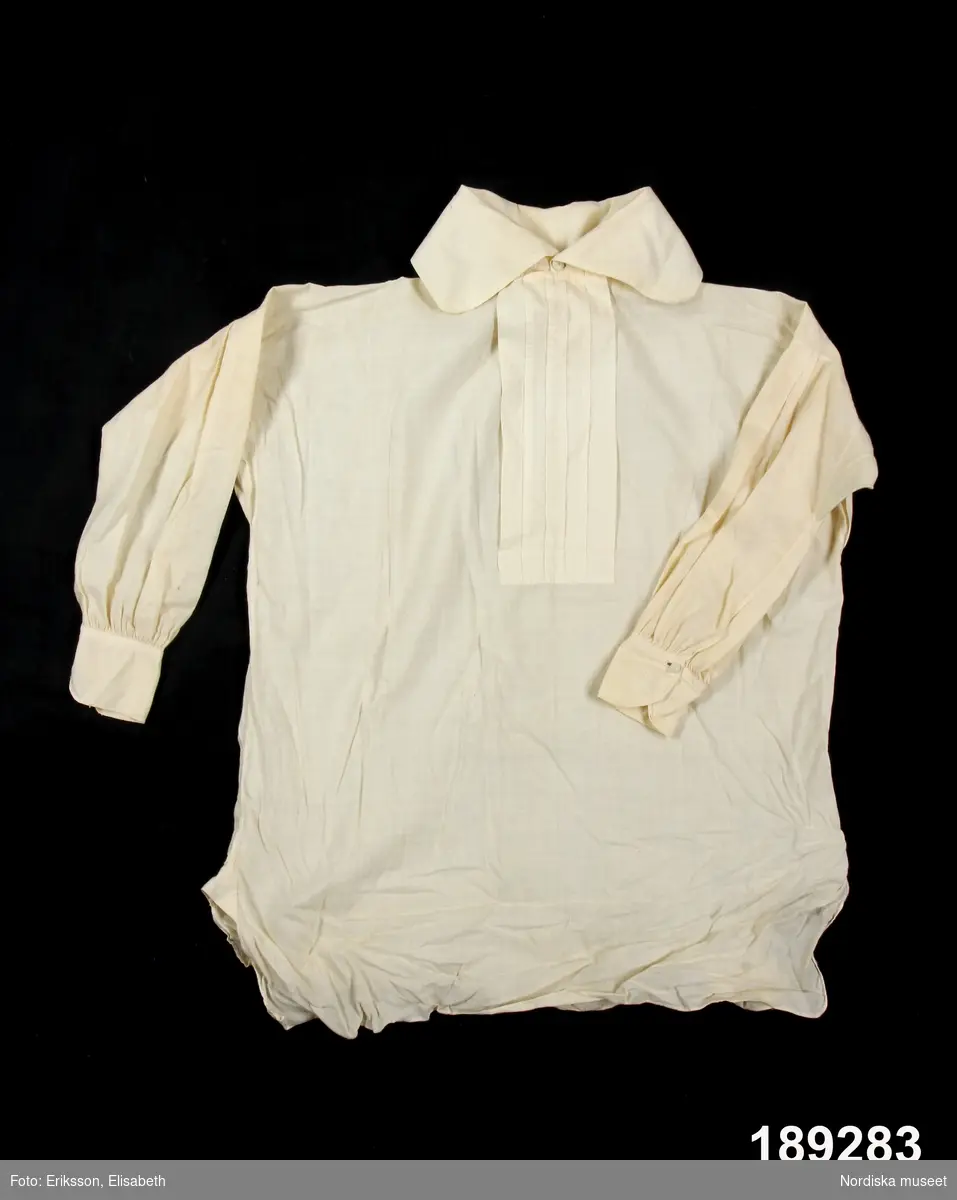 Skjorta i barnstorlek av vit tunn bomullslärft s.k. shirting. Bålen i ett stycke med sidsömmar, sidsprund med steglapp. Vidsydd rak ärm med  4 nedsydda veck på kullen,  ärmspjäll, ärmen rynkad mot 5 cm bred ärmlinning knäppt med liten trådknapp och knapphål. Hög rak krage som viks dubbel, knäpps i halsgropen med trådknapp och knapphål, halsspjäll med litet rynkparti, liksom mitt bak. Sprund fram  med förslag med 4 nedsydda veck på förslaget. Knäpps vid halsen med trådknapp och knapphål.  Skjortan sydd för hand med små fina stygn och fällsömmar .
Sydd 1867 av Johanna Jonsdotter f. 1817, givarens moder från Malmby i Närtuna.
/Berit Eldvik 2012-03-13