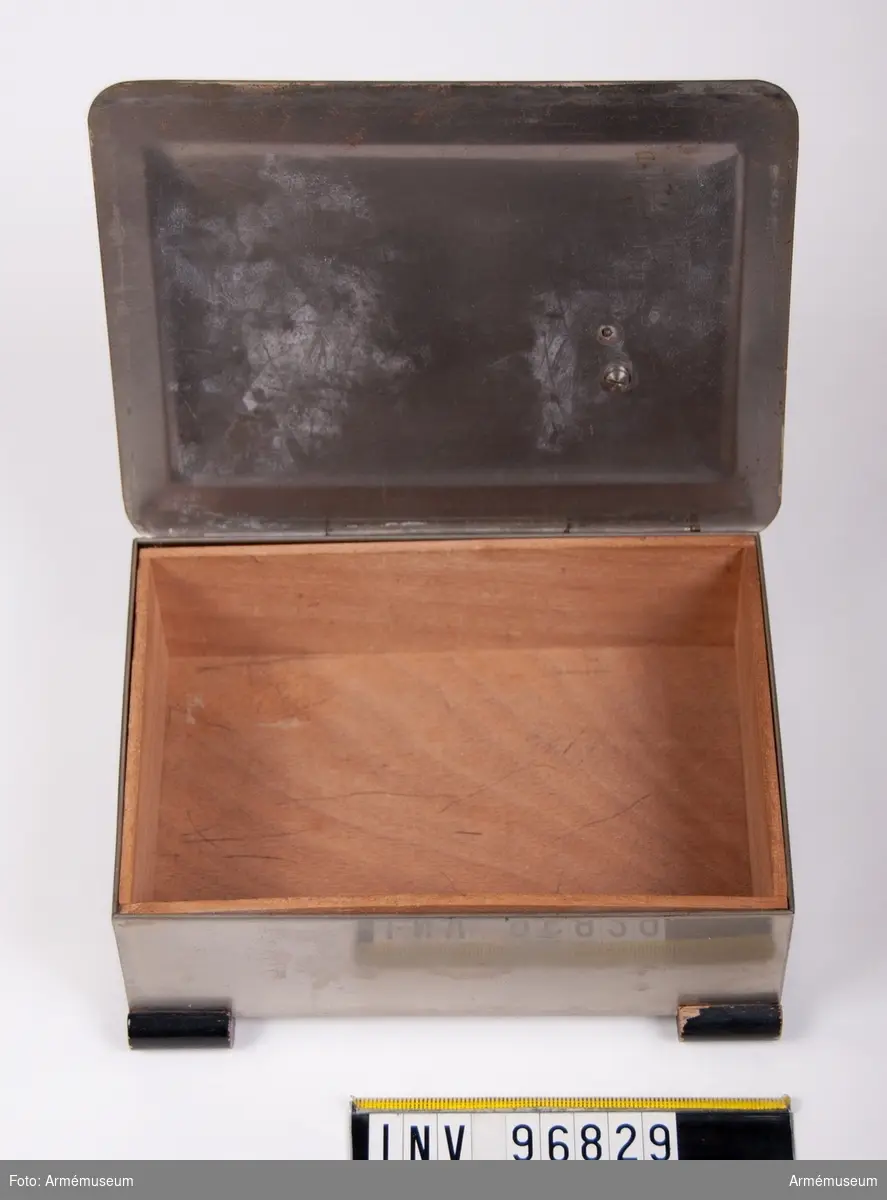 Skrin i metall med träinredning som möjligen har använts för att förvara cigarrer. Ovanpå locket en utsmyckning i form av en blomstergirlang med en tysk hjälm.