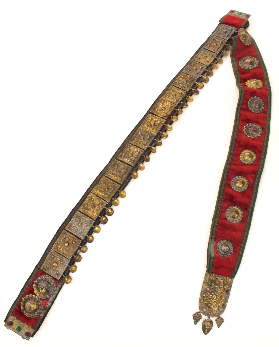 Belte med geheng. Av rød plysj, kantet med smal sort silkebord med små blomsterranker i grønt og fiolett. Beltet besatt med 16 kvadratiske utstansede forgylte sølvplater, hver med to nedhengende løv.  Flatene har rosettornamentikk.  Hektelås av forgylt blikk, de to deler hver med tre glasstener i rødt og grønt.  Ved siden av låsen på den ene side to runde forgylte beslag, utstanset arbeide, med ett løv i sentrum.  Festet til beltet et langt nedhengende geheng av samme materiale, besatt med ni runde utstansede forgylte beslag av forskjellig utførelse og størrelse.  Nederst beslått med avrundet endebeslag, forgylt, med utstanset ornamentikk på oversiden, et ansikt i midten.  Nederst tre løv. Beslaget har gullsmedmerker på baksiden (se Påført tekst) 
Beltet er foret med rødt bomullsstoff med sorte prikker.  Gehenget foret med sort silkebånd, blomsterranke i rosa, og tunget rosa kant.