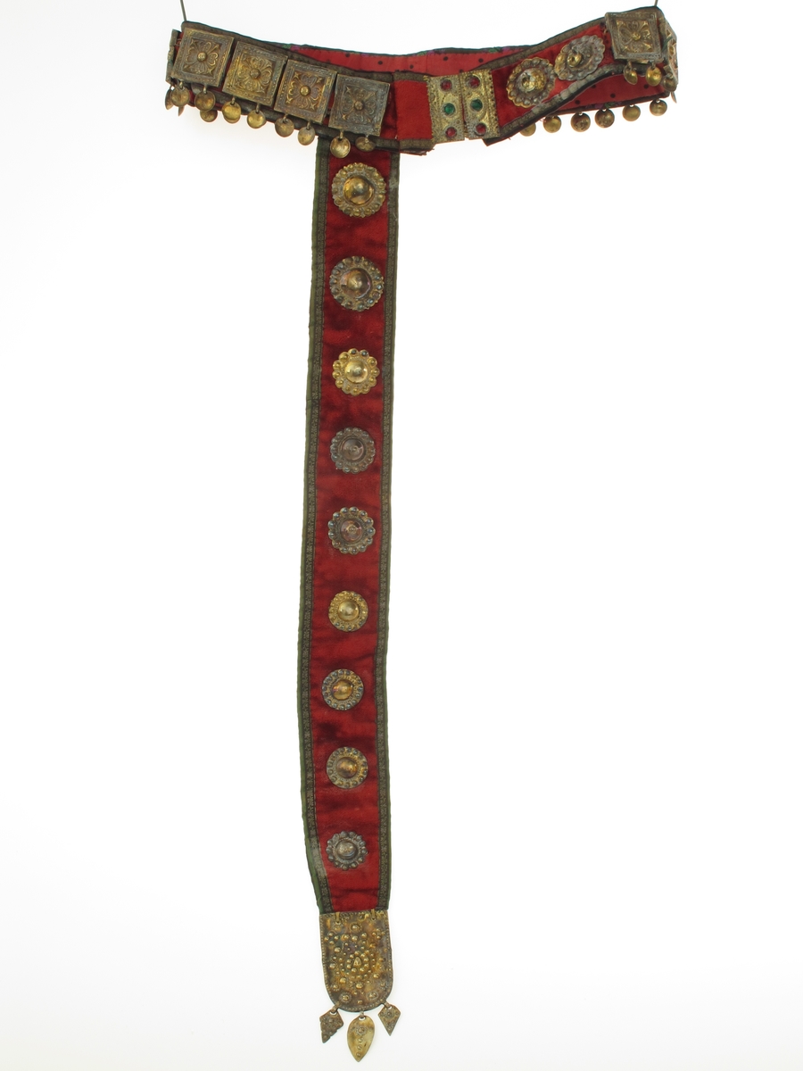 Belte med geheng. Av rød plysj, kantet med smal sort silkebord med små blomsterranker i grønt og fiolett. Beltet besatt med 16 kvadratiske utstansede forgylte sølvplater, hver med to nedhengende løv.  Flatene har rosettornamentikk.  Hektelås av forgylt blikk, de to deler hver med tre glasstener i rødt og grønt.  Ved siden av låsen på den ene side to runde forgylte beslag, utstanset arbeide, med ett løv i sentrum.  Festet til beltet et langt nedhengende geheng av samme materiale, besatt med ni runde utstansede forgylte beslag av forskjellig utførelse og størrelse.  Nederst beslått med avrundet endebeslag, forgylt, med utstanset ornamentikk på oversiden, et ansikt i midten.  Nederst tre løv. Beslaget har gullsmedmerker på baksiden (se Påført tekst) 
Beltet er foret med rødt bomullsstoff med sorte prikker.  Gehenget foret med sort silkebånd, blomsterranke i rosa, og tunget rosa kant.