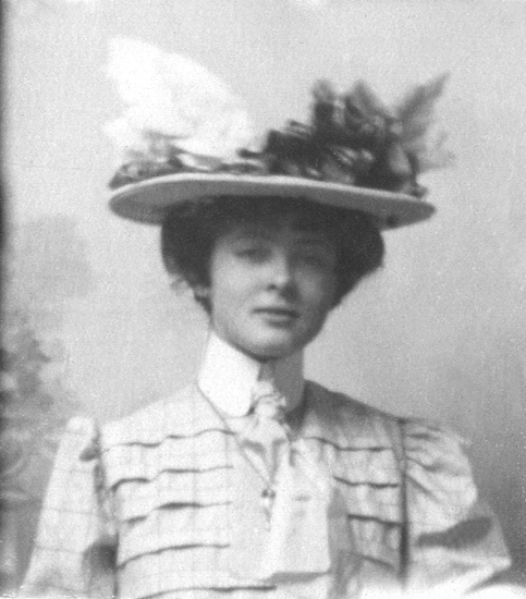 En ung dam, iklädd ljus blus med hög krage m.m.
På huvudet en hatt med stora dekorationer.
Något suddigt foto.