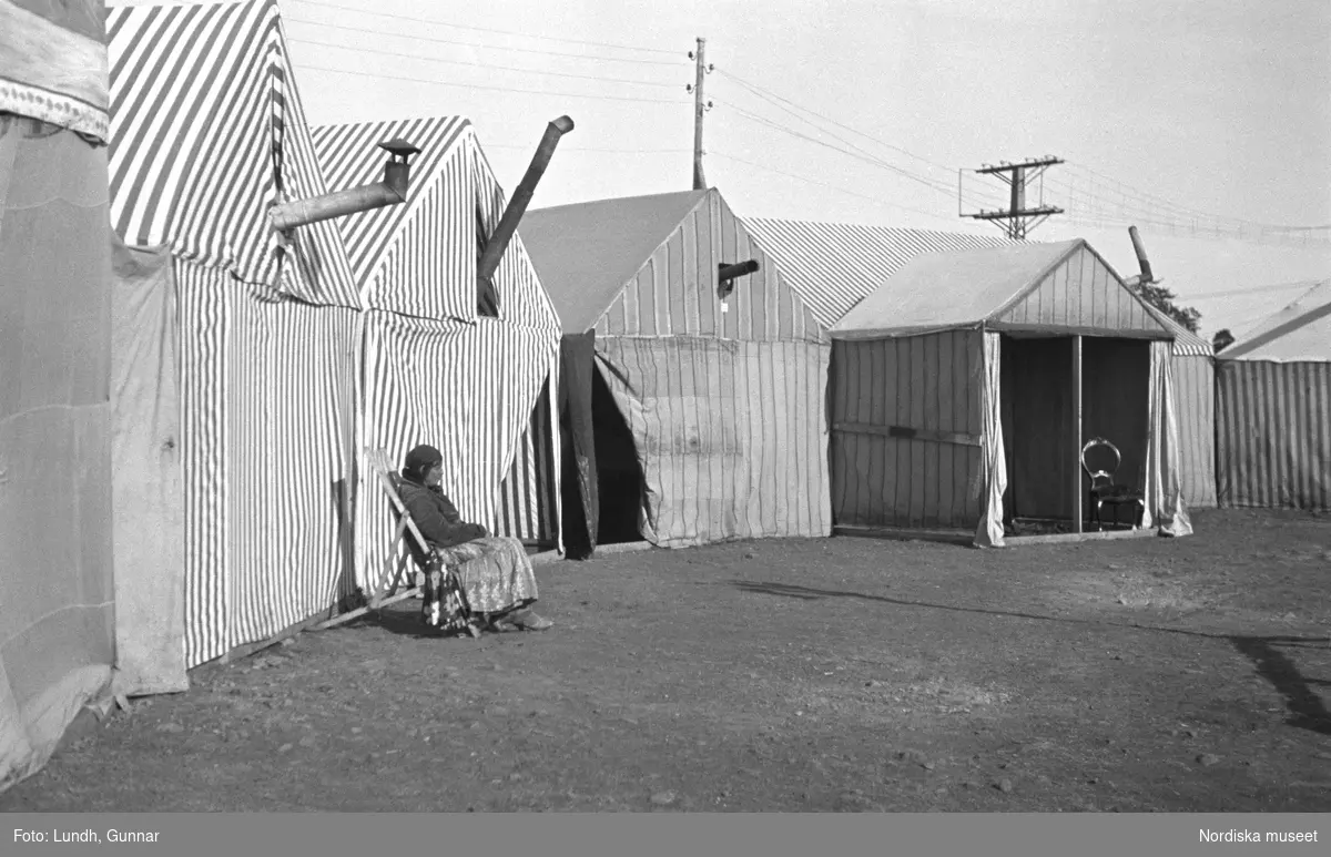 En ensam kvinna sitter i en stol framför en rad tält. Familjen Taikons läger i Johanneshov, södra Stockholm.