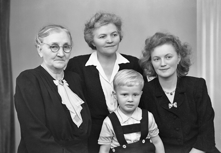 Foto av fyra generationer. Tre kvinnor i olika åldrar och en pojke i hängslebyxa. 
Ateljéfoto.