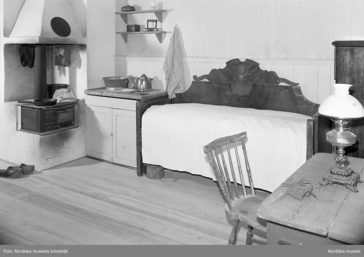 Interiör från arbetarbostaden i block 4 på Nordiska museets jubileumsutställning "Svenskens 100 år - känn dig själv". I köket med vedspis och lavoar står även en högt uppbäddad utdragssäng och ett köksbord med pinnstol. På bordet står en fotogenlanmpa.