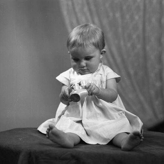 Foto av en liten flicka i klänning som sitter på ett tygklätt bord.
Helfigur. Ateljéfoto.