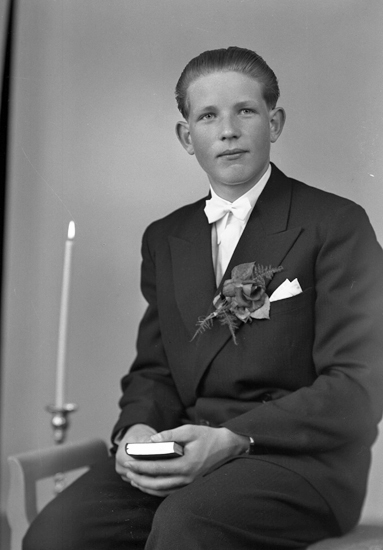 Foto av en ung man i konfirmationskostym. Bredvid honom syns en tänd golvljusstake.
Knäbild. Ateljéfoto.
