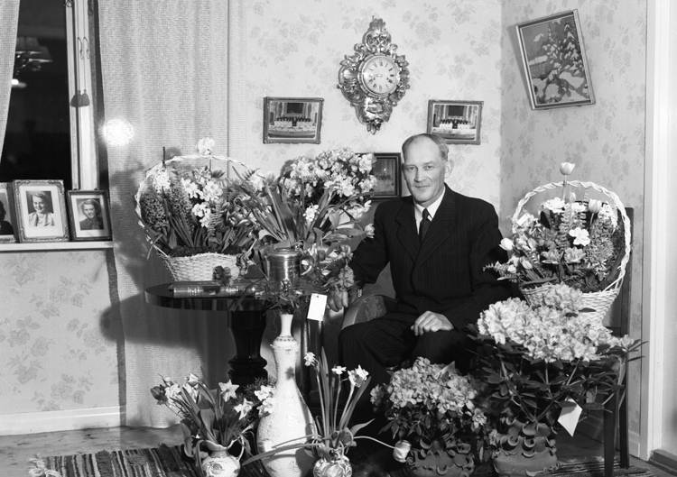 Foto av en man i kostym som sitter i ett vardagsrum. Han är omgiven av mängder med blommor.