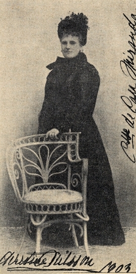 Foto på Christina Nilsson. Hon står bakom en korgstol. 
Hon bär svart klänning (sorg- ?) och hatt.