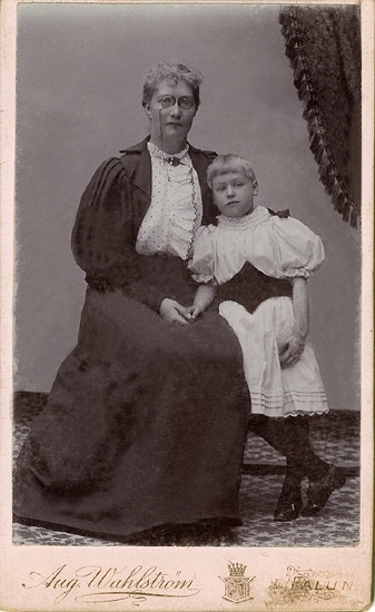 Grupporträtt (helfigur) av en sittande kvinna med pincené, iklädd mörk klänning med prickig blus, med en flicka i vit klänning med mörkt skärp och svarta strumpor, stående bredvid sig.