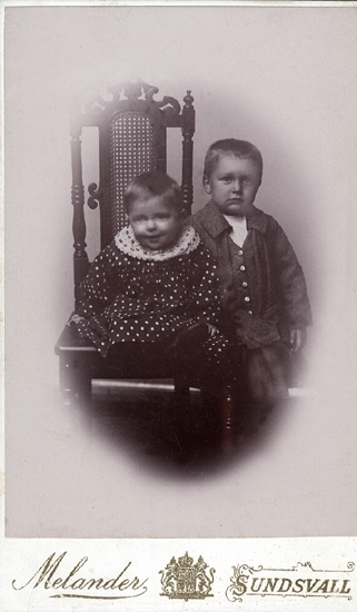 Porträtt (helfigur) av två små barn, en liten flicka i prickig kolt (?) som sitter på en högryggad stol. 
Bredvid stolen (till höger) står en liten pojke i jacka och knäbyxor.
