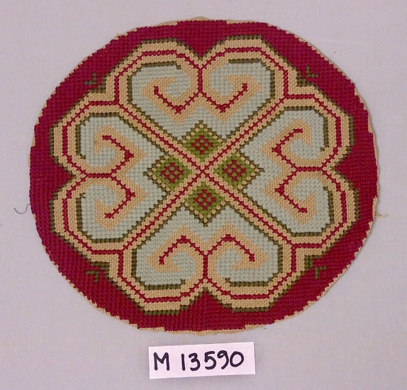 Broderi till kudde eller möjligen pall, på
säckvävsliknande material. En rund oval med 
måtten: 355 x 335 mm.
Ett geometriskt mönster i korsstygn och förskjutna korsstygn
i färgerna rött, ljusblått och grönt. 
Inskrivet i huvudkatalog 1944.