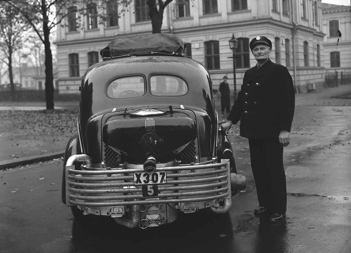 En chaufför, droskägare P J Green, står på Teaterplan framför Teatern i Gävle vid en X307 förlängd 1937  Dodge D-5 Touring Sedan med gengasaggregat. I bakgrunden Norra skolan.