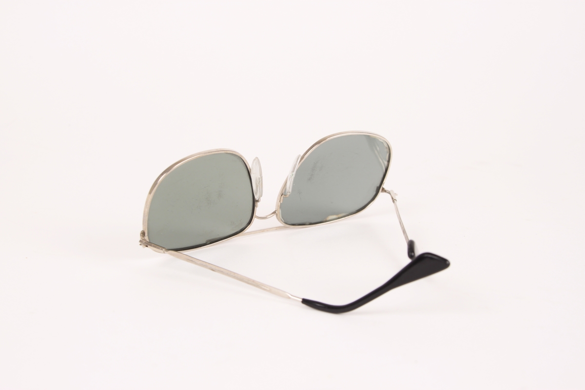 Et par solbriller med metallinnfatning. Brillens modell er pilotbrille, med sølvfarget innfatning og svarte glass. Svart plast på enden av hver brillestang.