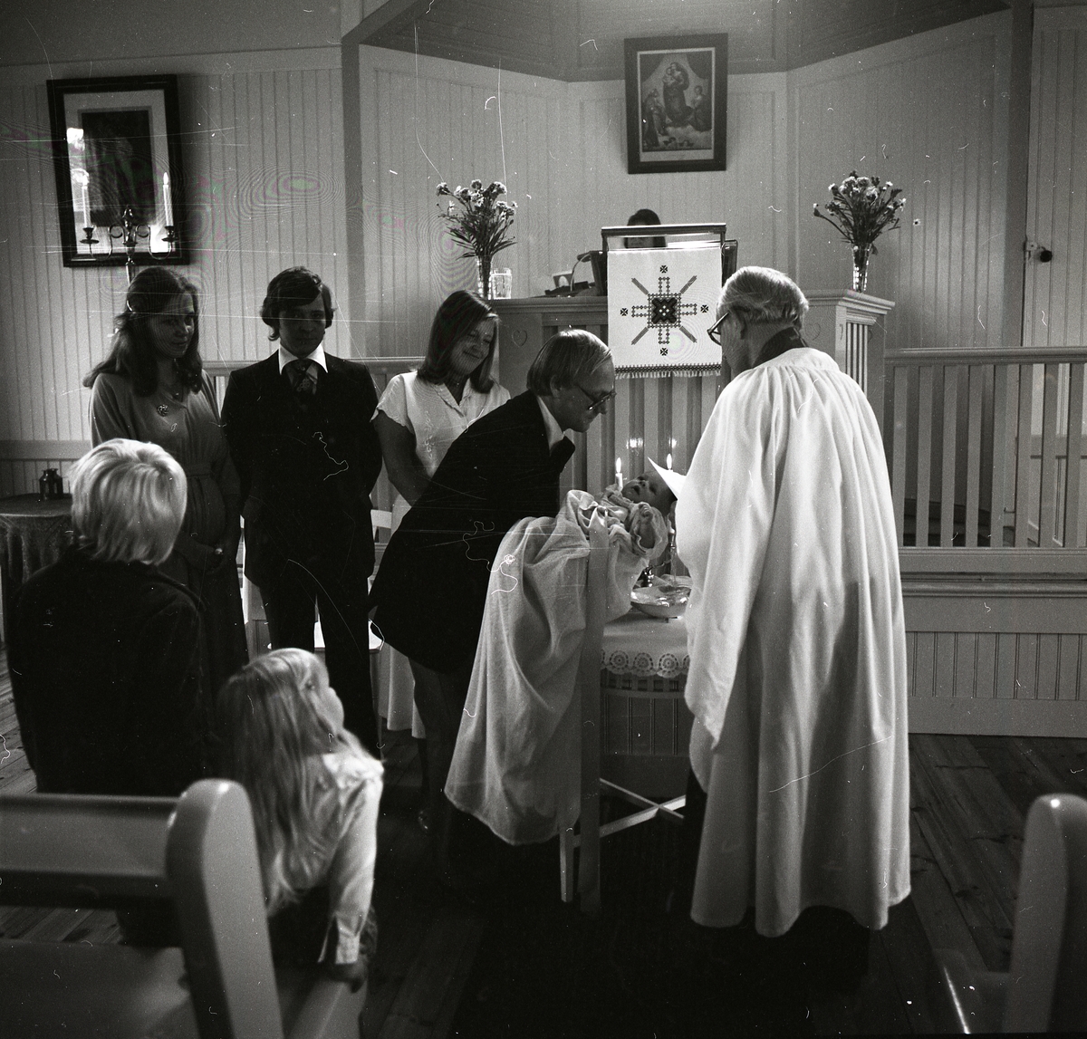 I kyrkans bänkrader sitter två barn och tittar omkring sig. De verkar måttligt intresserade av dopet som utspelar sig framför dem. Prästen iklädd vit kappa står framför bordet med vigvatten i en metallskål. En man sänker försiktigt ned barnen mot skålen för att döpas. Barnet bär en lång dopklänning och bakom dess huvud lyser två ljus. Två kvinnor och en man beskådar händelsen med glada miner.