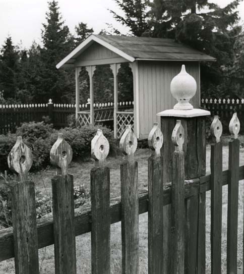 En trädgård bakom ett staket. I trädgården finns en paviljong smyckad med snickarglädje.