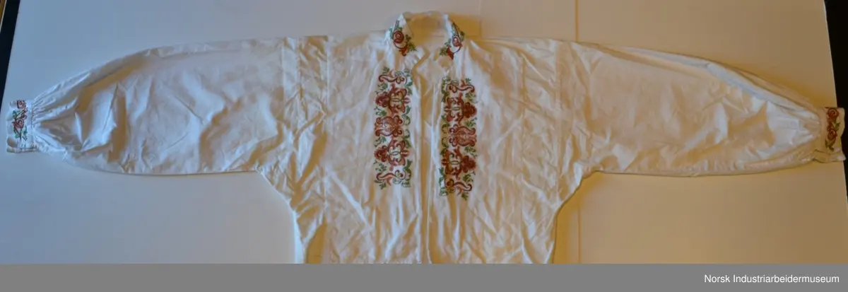 Plattsøm på kraven, mansjettene og bringa. Skjorten er åpen foran og helt ned.