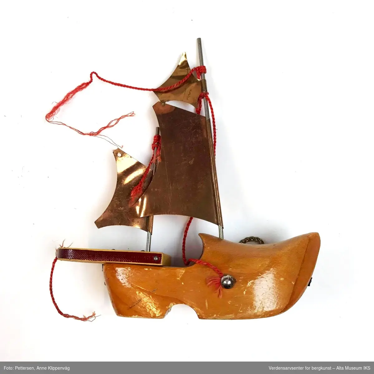 Form: Treskoform (venstre) med påsatt akterdel og to master med med seil av metall. Turistsuvenir fra Hammerfest.
Trehvitt skrog med røde og svarte striper, seil i gullmetall-folie, rødt tauverk.