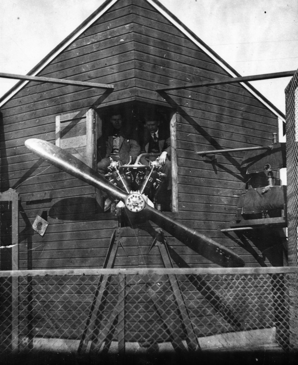 Flygmotor placerad i motorprovbock på flygverkstäderna i Malmslätt. Två män sitter i huset bakom motorn.