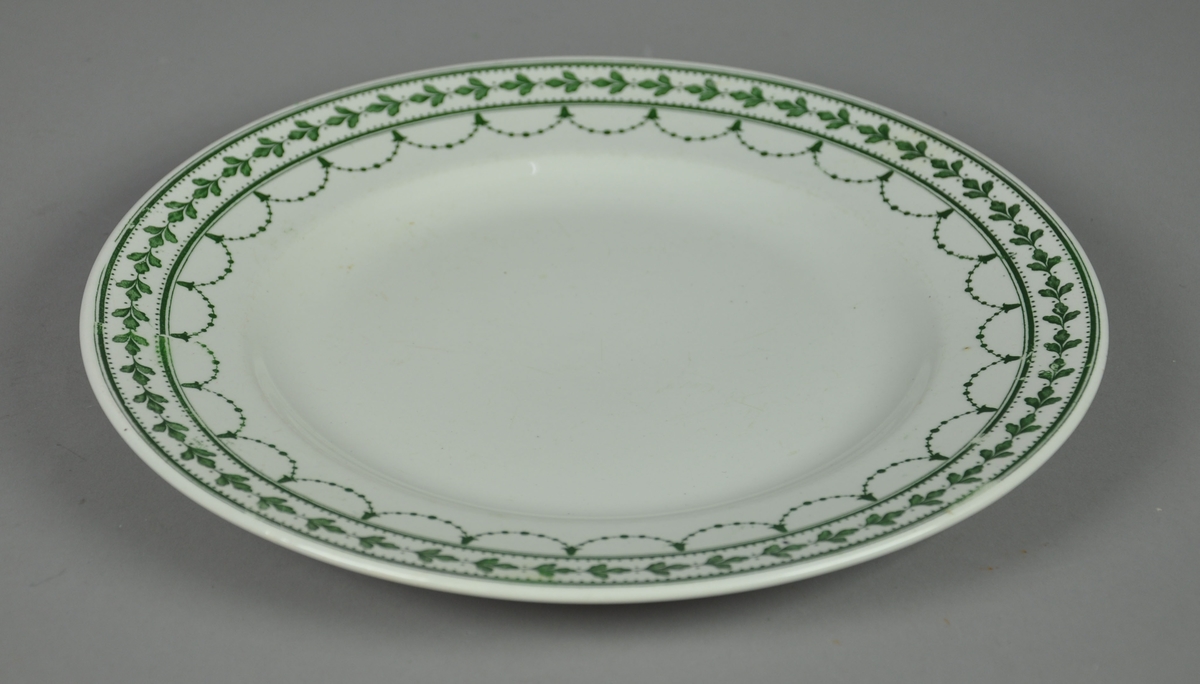 Rund tallerken av keramikk, steingods, med grønn bord. Borden består av eikekrans og guirlandere.
Mønsteret heter Fontainebleau.