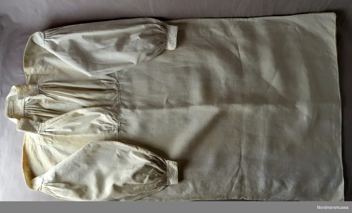 Lang herreskjorte med labbesøm på halslinning og ermlinning. Sydd saman av to stykke. Øverste stykket har broderi og legg. (33 cm).  Ingen knepping.