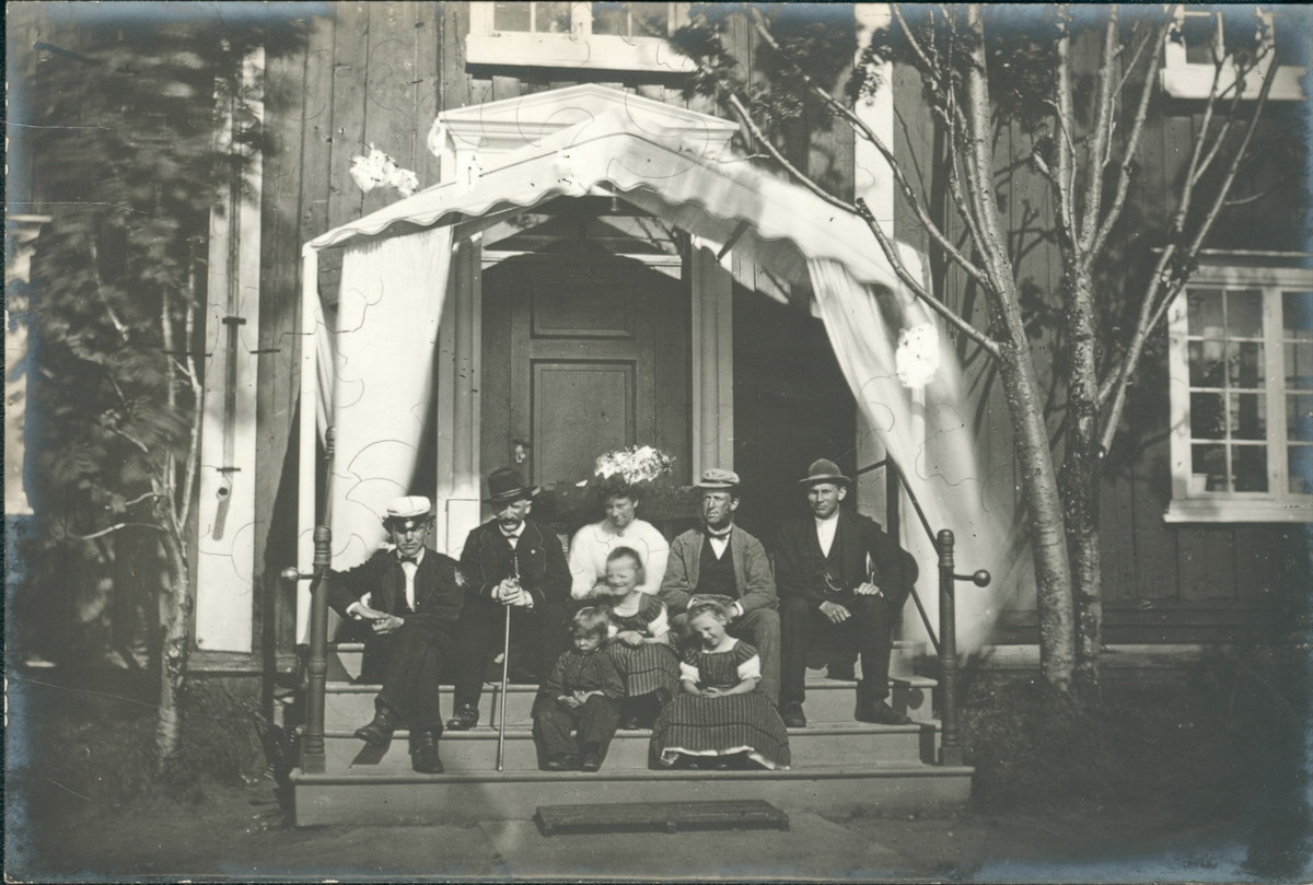 Gruppefoto.  Meyerfamilien samlet på Meyergårdens trapp. Med tre ukjente menn. Handelsmann L. A. Meyer med hatt og stokk ( nummer to fra venstre) og hans tre barn og kone nummer to Hanna. Hvit baldakin over døra. Sannsynligvis tatt rundt 1870.