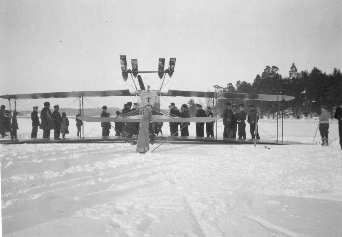 Ett havererat militärt propellerflygplan med skidor ligger upp och ned på isen. I bakgrunden står män ur flottan.