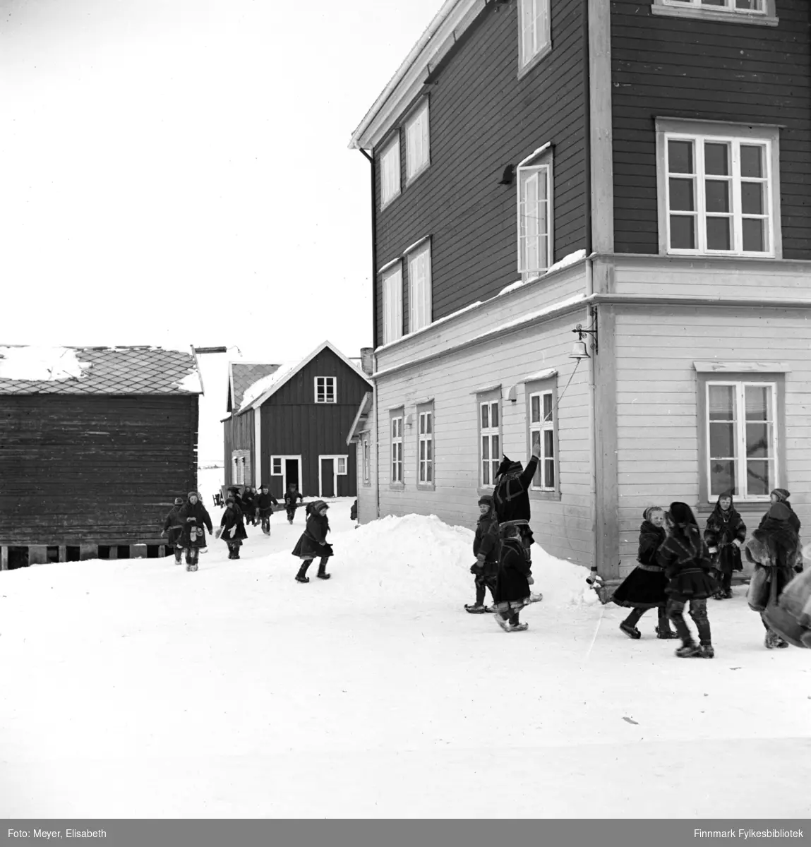 Barn leker ute i skolegården utenfor Kautokeino skoleinternat. En lærer står og ringer med skoleklokka som henger på hjørnet av bygningen. Skolen ble bygd i 1906. Påbygd i 1932. Skolen ble brent i 1944.