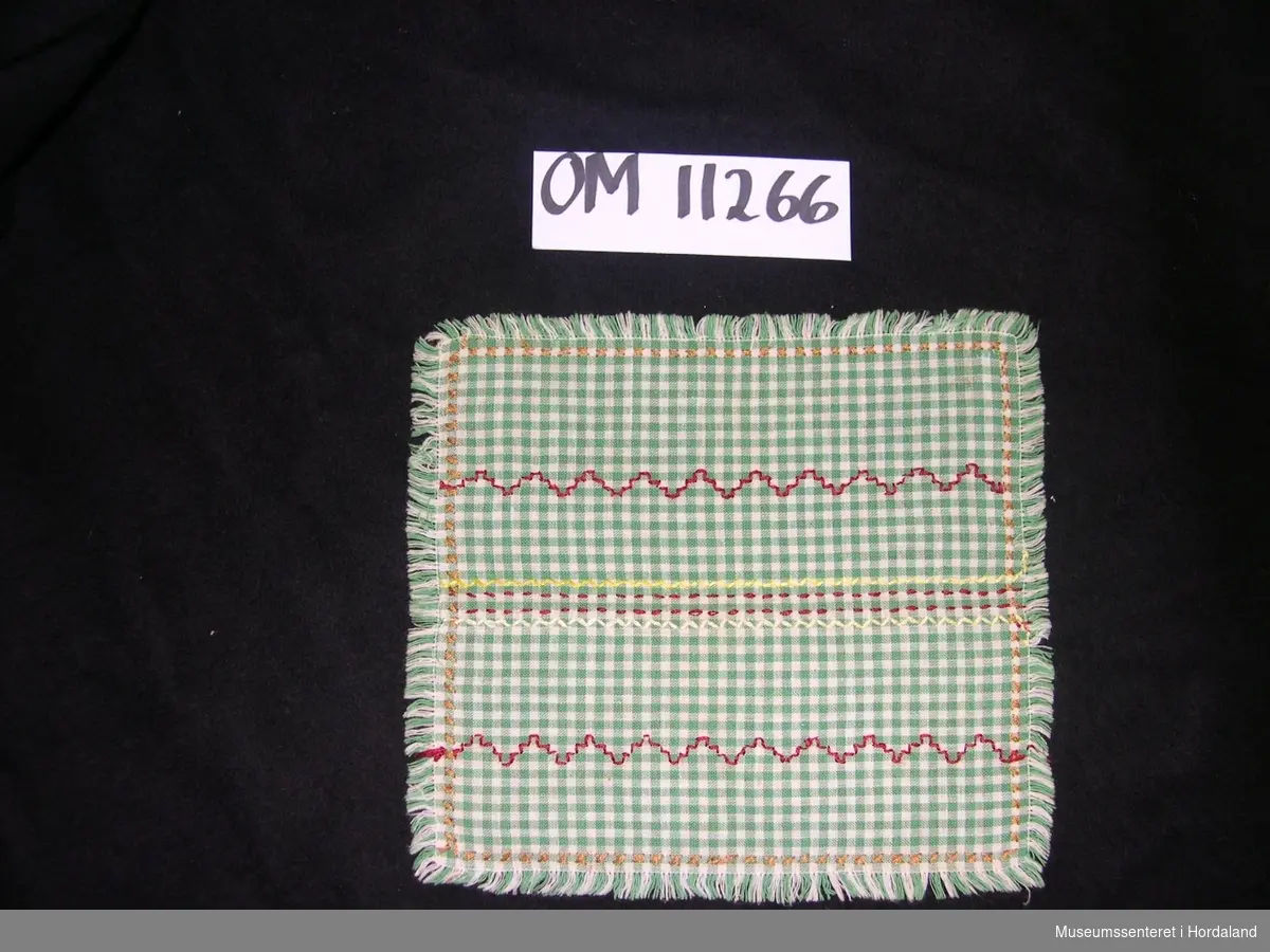 Kvadratisk brikke i grønt- og hvitrutete stoff med sydd sikksakk-mønster og korssting i rødt, hvitt, gult og brunt.