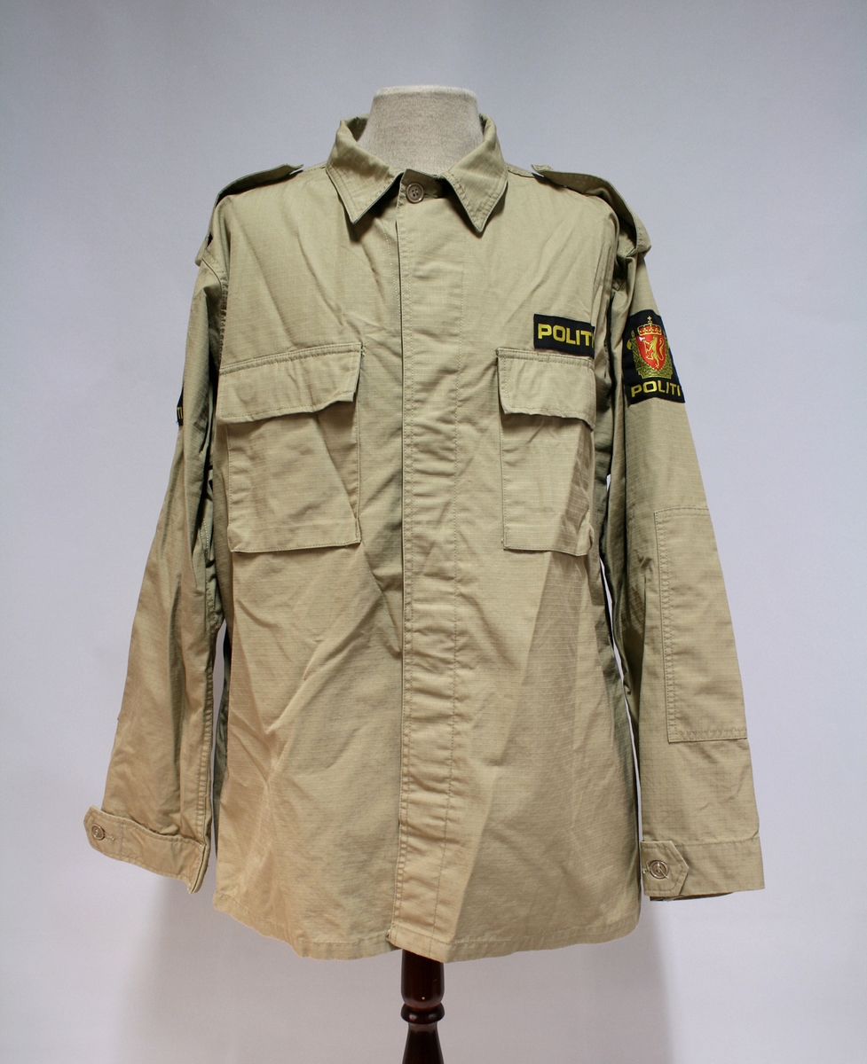 Khakifarget jakke med politimerking etter 1995-reglementet. To brystlommer. Plastknapper. Ingen distinksjoner.