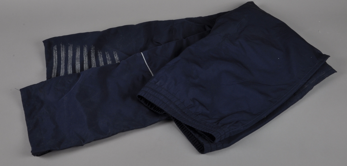 Blå overtrekksbukse med logo for UMBRO. Buksen har glidelås nederst ved hver ben, og refleksstriper på venstre ben. Buksestørrelsen er S.