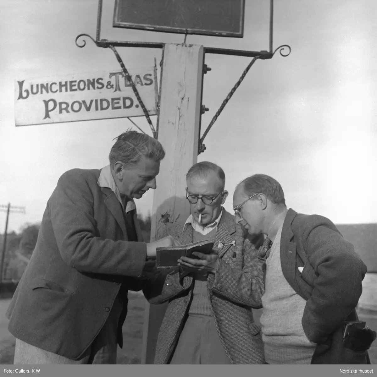 Tre män tittar intresserat i en bok framför en skylt.