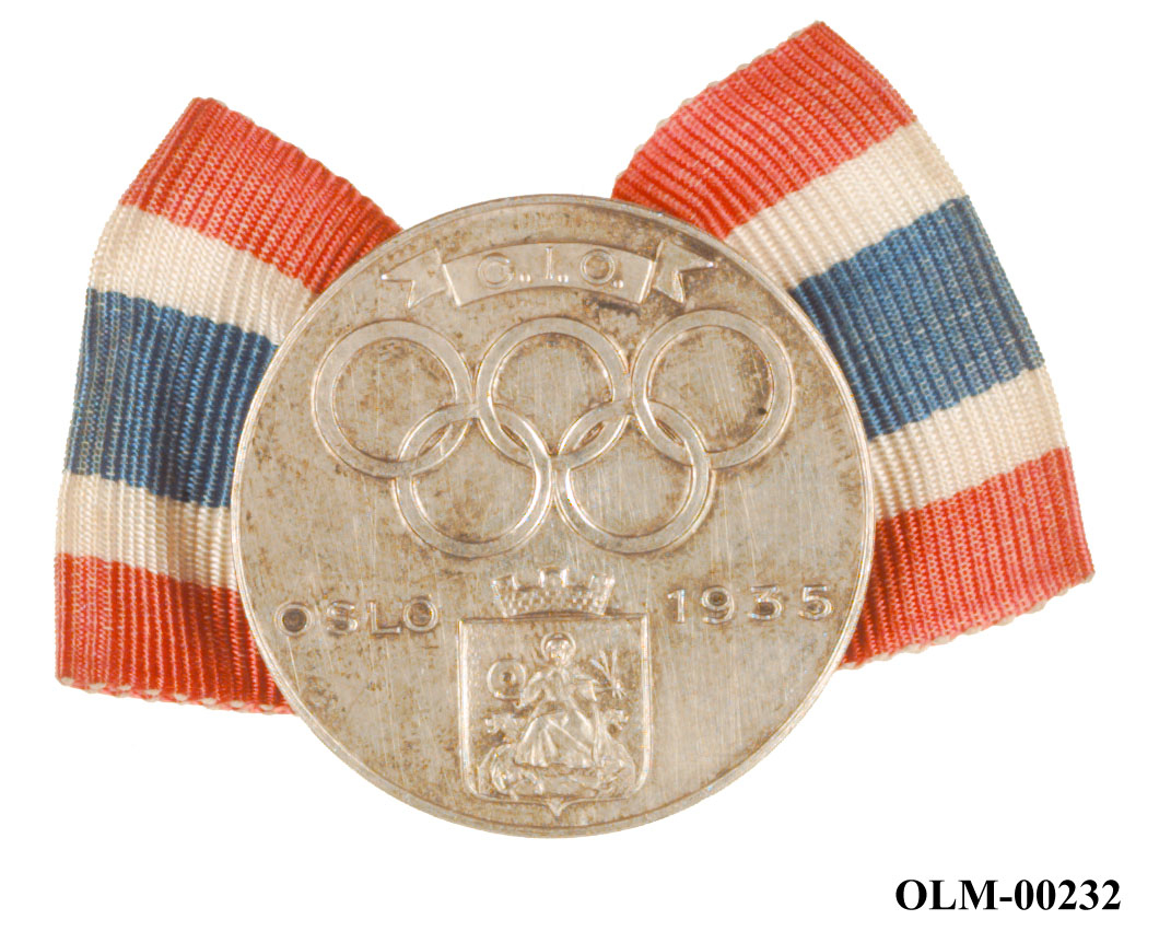 Sølvfarget merke med Oslos byåpen St. Hallvard, innskriften Oslo 1935, de olympiske ringene og C.I.O. Merket er festet på et bånd med det norske flaggs farger.