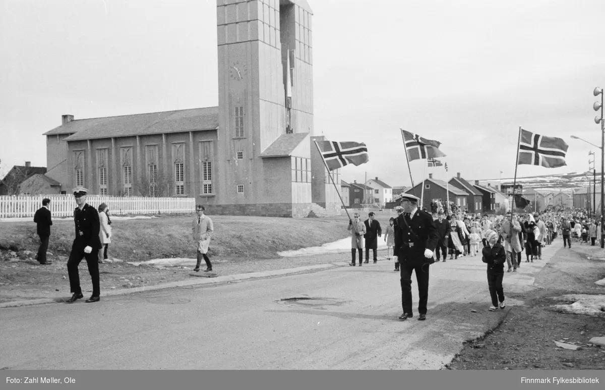 Vadsø, 17.mai 1970. Korpset spiller i gatene. Vadsø kirke i bakgrunnen. Politiet fremst i toget.
